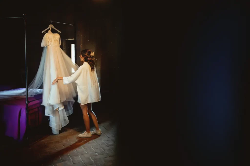 The bride admires her wedding dress on a hanger in a semi-dark room.