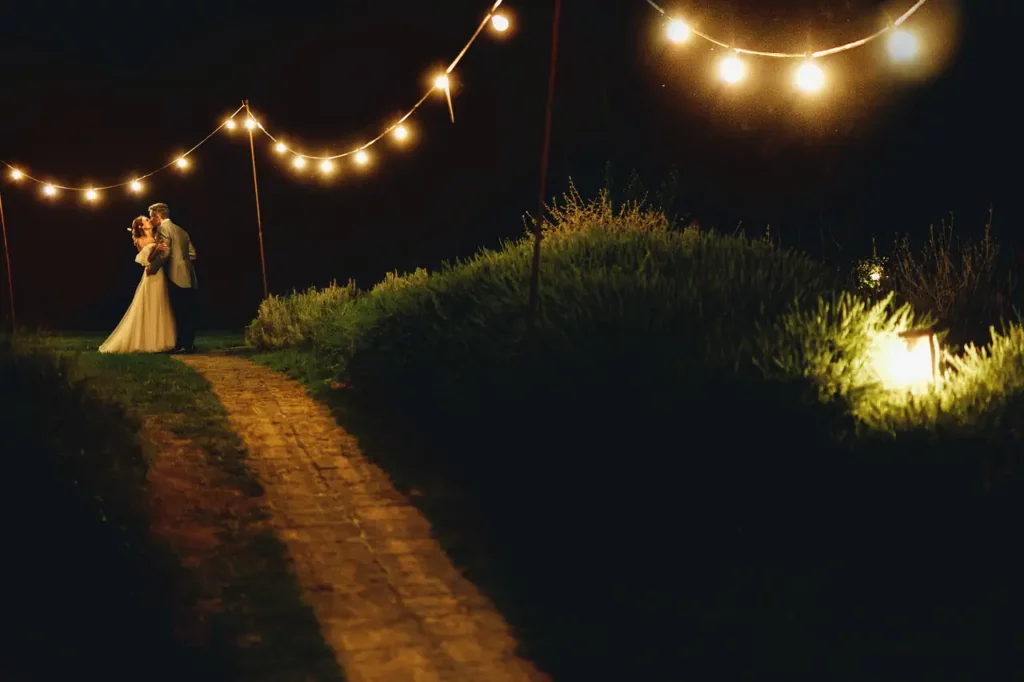 The bride and groom, standing, kiss under a row of lights, at night.