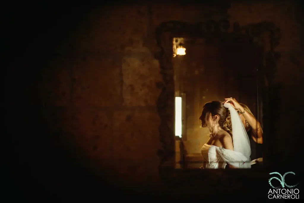 La sposa si aggiusta il velo davanti allo specchio con l'aiuto della parrucchiera.