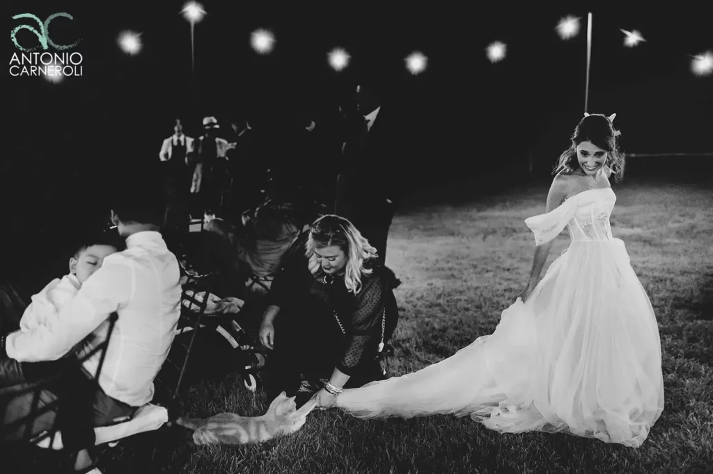 The bride, in a black and white photo, and a guest kneeling on the grass clearing the bride's dress of a kitten