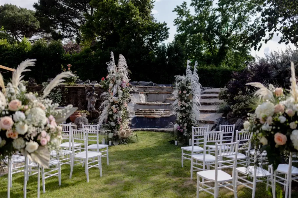 Outdoor ceremony setup with white chairs and floral arrangements leading to an altar.