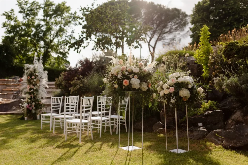 Outdoor wedding ceremony setup with white chairs and flower arrangements on a sunny day.