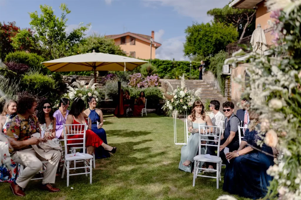 Guests seated at an outdoor ceremony with floral decorations on a sunny day.