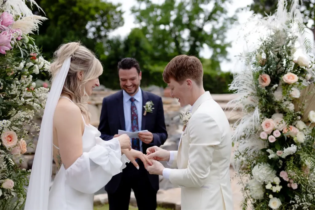 A couple exchanges rings during an outdoor wedding ceremony, with a smiling officiant in the background.