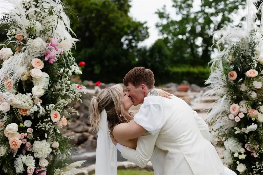 A couple kissing during the wedding ceremony, framed by a floral arch.