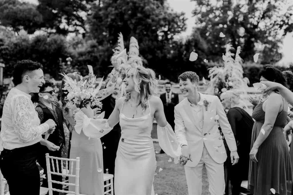 Smiling newlywed couple as they walk hand in hand, with guests throwing flower petals to celebrate their wedding.