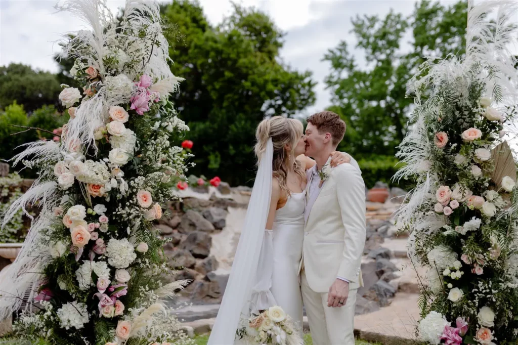 A couple kissing during the wedding ceremony, framed by floral arrangements.