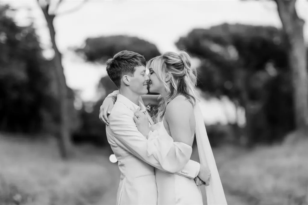 A monochrome image of a smiling couple in wedding dresses, hugging and gazing into each other's eyes outdoors.