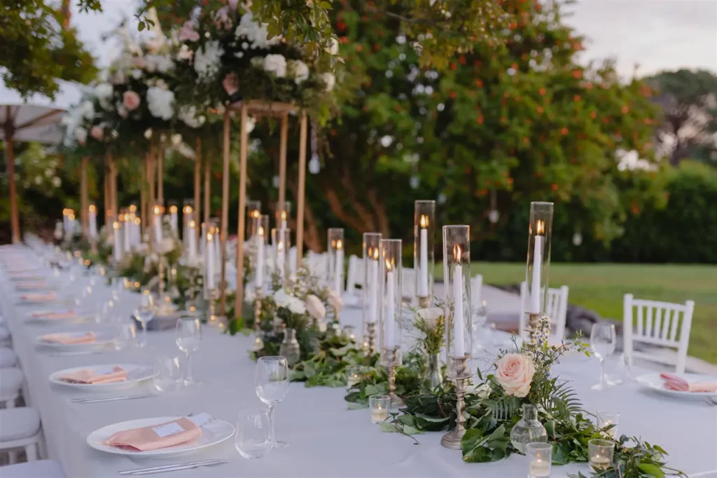 Outdoor wedding reception table elegantly set up with flower arrangements and candles lit at dusk.