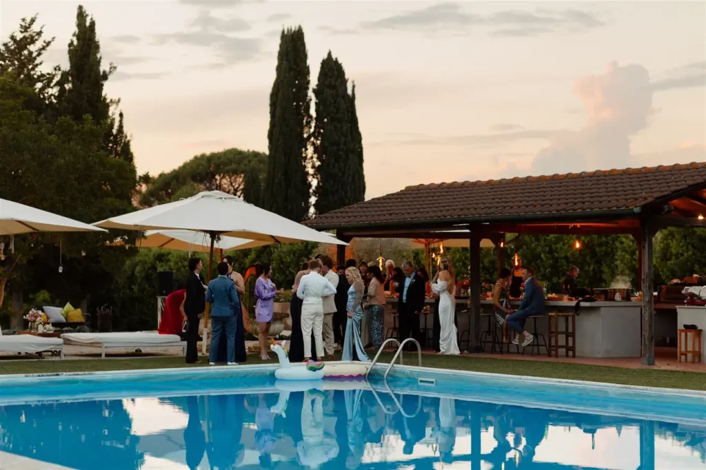 An outdoor evening reception by the pool with guests socializing under a canopy and umbrellas.