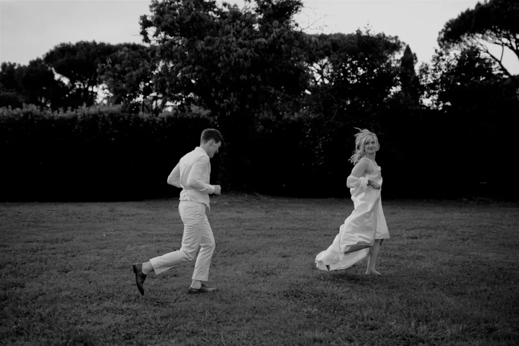 A groom chases the bride through a grassy area.
