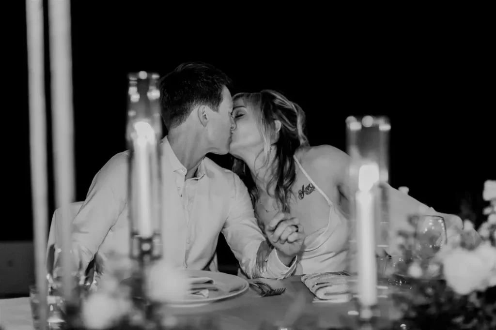 A black-and-white photo of the bride and groom kissing in front of a table set with candles during a reception.