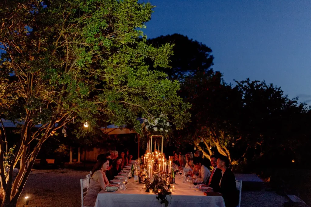 An outdoor evening reception with guests seated around a table adorned with candles under the trees.