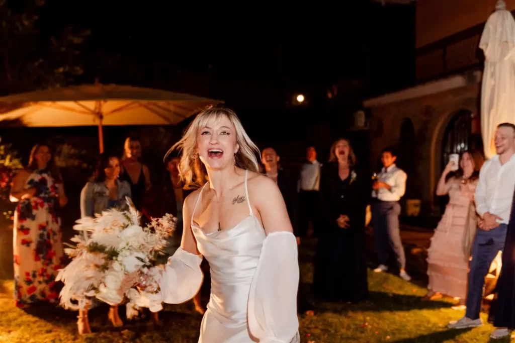 A bride prepares to throw her bouquet with guests in the background during an evening wedding ceremony.
