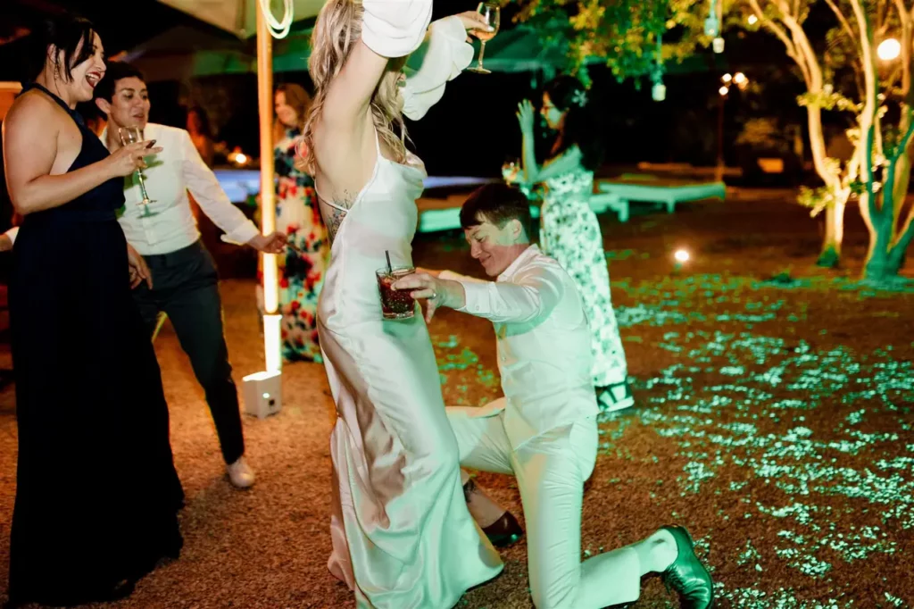 The bride in a white dress is dancing with the groom on her knees, both holding drinks in their hands.