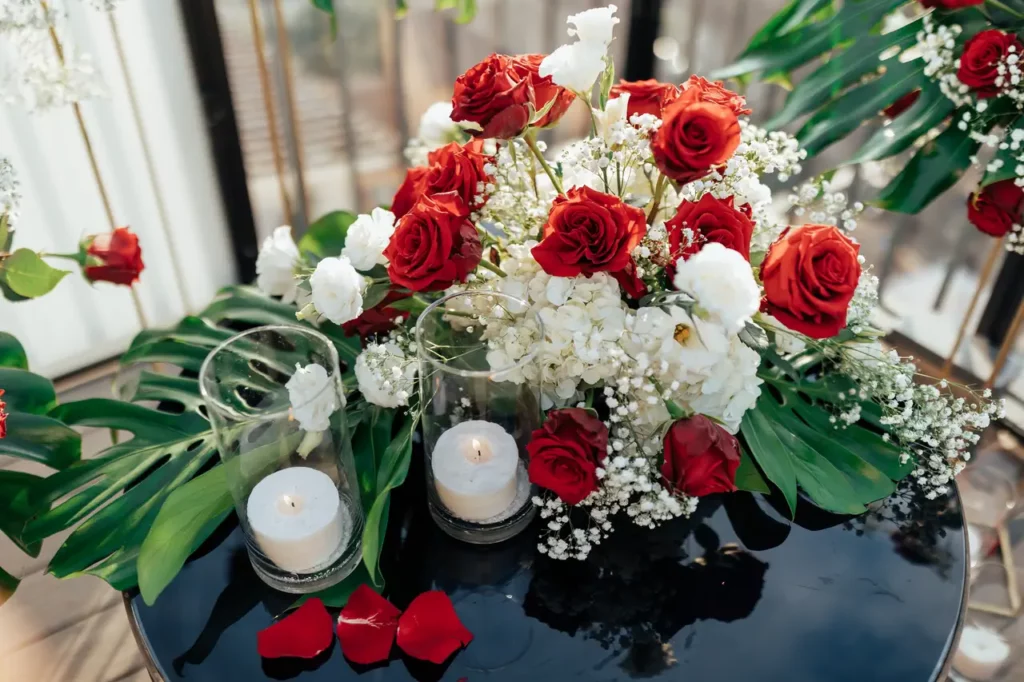 Un centrotavola floreale con rose rosse, fiori bianchi e foglie verdi, accompagnato da candele accese su un tavolo riflettente.