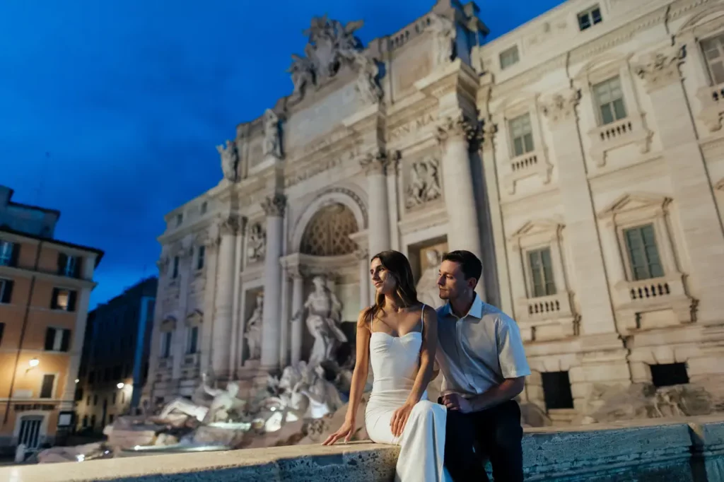A couple sits together in front of the Trevi Fountain in Rome at dusk.