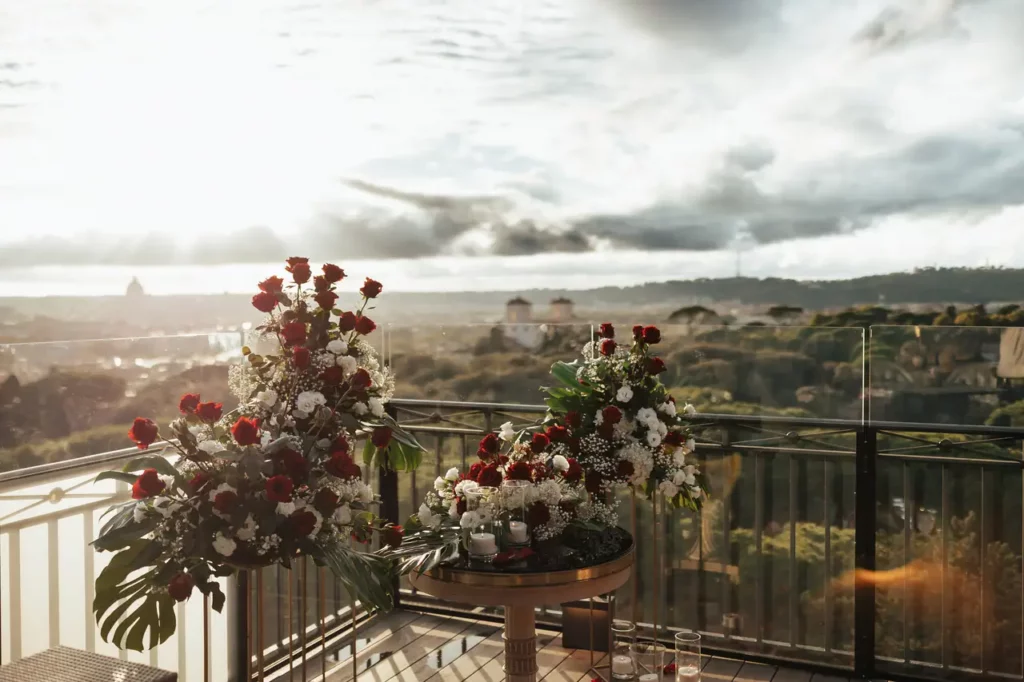 A rooftop terrace overlooking Rome with floral arrangements in the foreground under a sky with clouds.