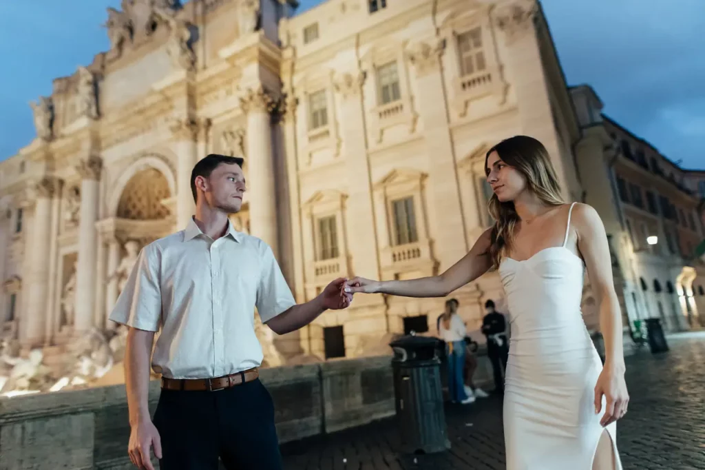 A couple holding hands in front of the Trevi Fountain at dawn.