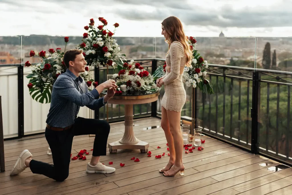Un uomo in ginocchio fa una proposta di matrimonio ad una donna su un terrazza decorato con fiori e petali di rosa.