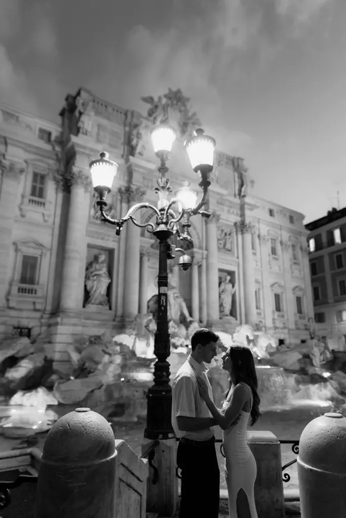 Couple embracing near an illuminated streetlight with the trevi fountain in the background at night.