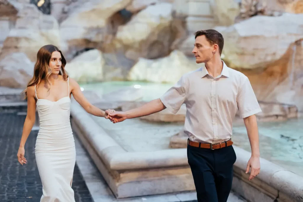 A couple holding hands as they walk past the Trevi Fountain.