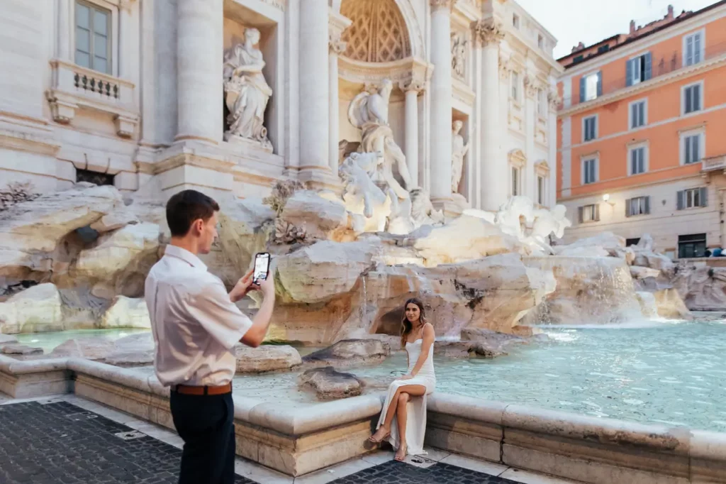 A man takes a picture of a woman sitting next to the Trevi Fountain in Rome.
