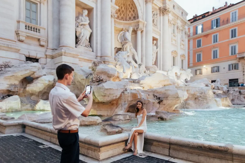 A man takes a picture of a woman sitting next to the trevi fountain in rome.