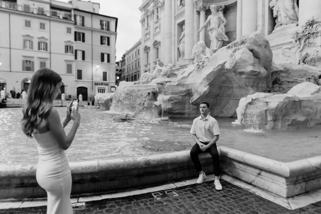A woman takes a picture of a man sitting next to the Trevi Fountain.