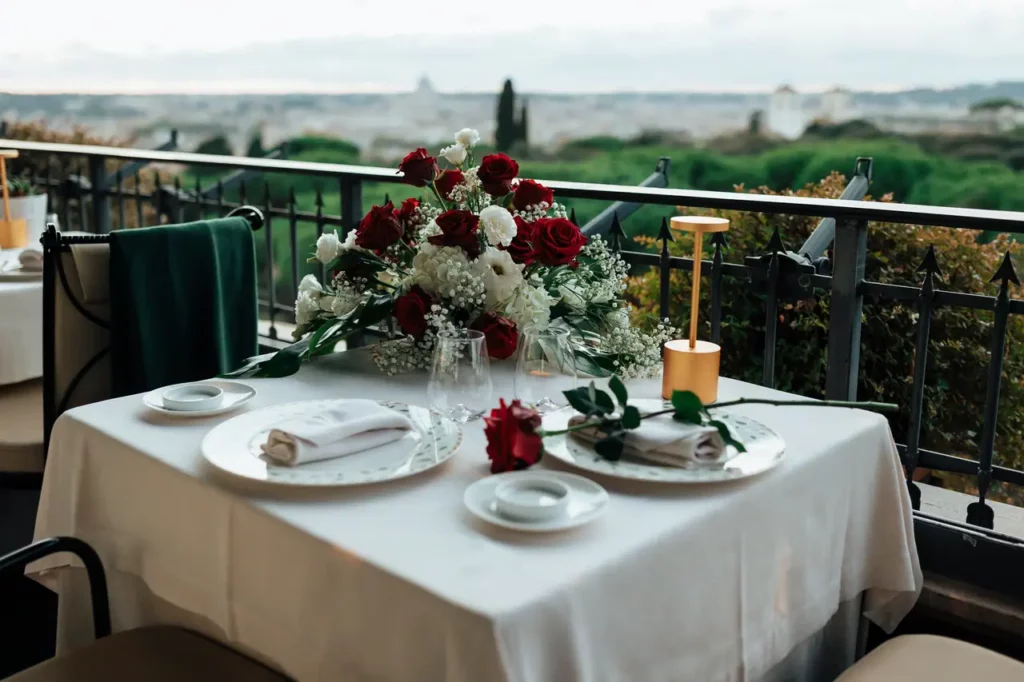 Elegante tavolo da pranzo da esterno apparecchiato per due con un mazzo di rose rosse e vista sul paesaggio di Roma al tramonto.