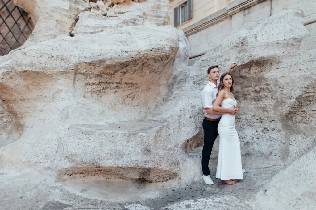 A couple in formal attire pose together against the stone backdrop of the trevi fountain.