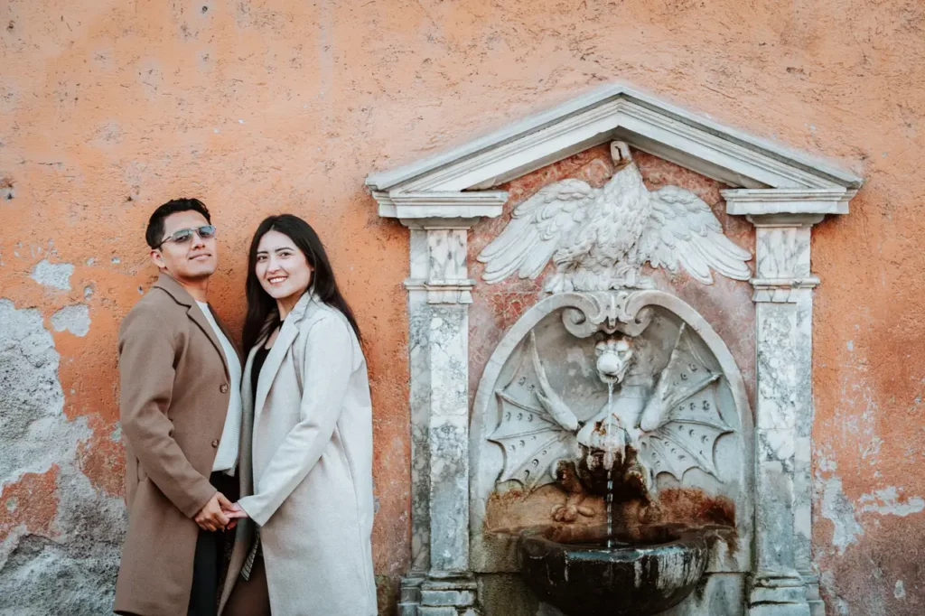 A couple stands, holding hands and smiling, in front of a decorative stone fountain set against a textured orange wall.