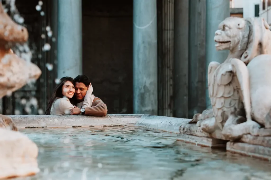 A couple leans on the edge of the pantheon fountain, smiling and embracing, with columns and stone statues visible in the background.