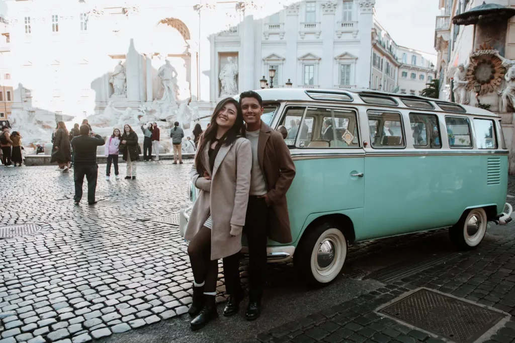 A couple is standing next to a vintage turquoise van on a cobblestone street, with historic buildings and a crowd of people moving in the background near the trevi fountain.
