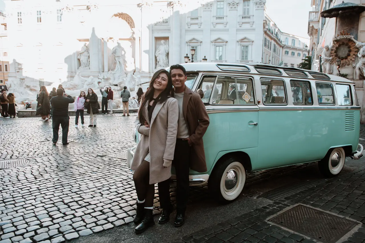 A couple is standing next to a vintage turquoise van on a cobblestone street, with historic buildings and a crowd of people moving in the background near the Trevi Fountain.