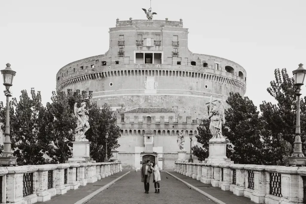 A couple walks hand in hand across a bridge toward the historic castel sant'angelo in rome, italy. The black-and-white image elegantly captures the statues that line the bridge.