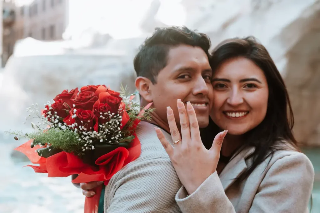 A smiling couple poses in front of a stone structure; the woman shows an engagement ring holding a bouquet of red roses wrapped in red paper.