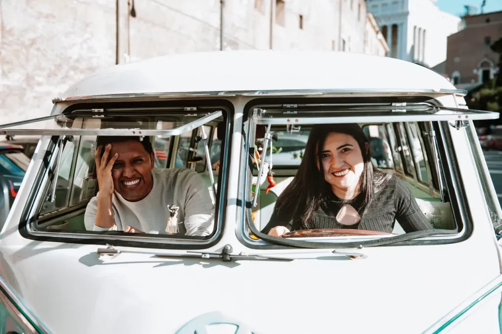 A man and a woman smiling and waving from the front seats of a vintage van.