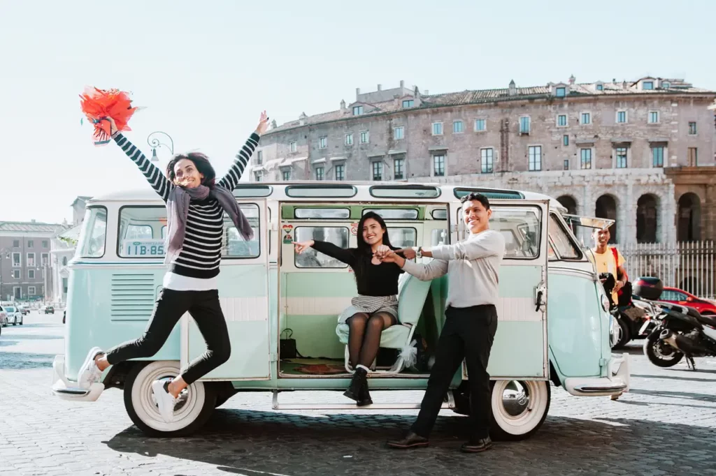 A couple poses near a vintage van in a sunny square with simona celani jumping with a bouquet of flowers.