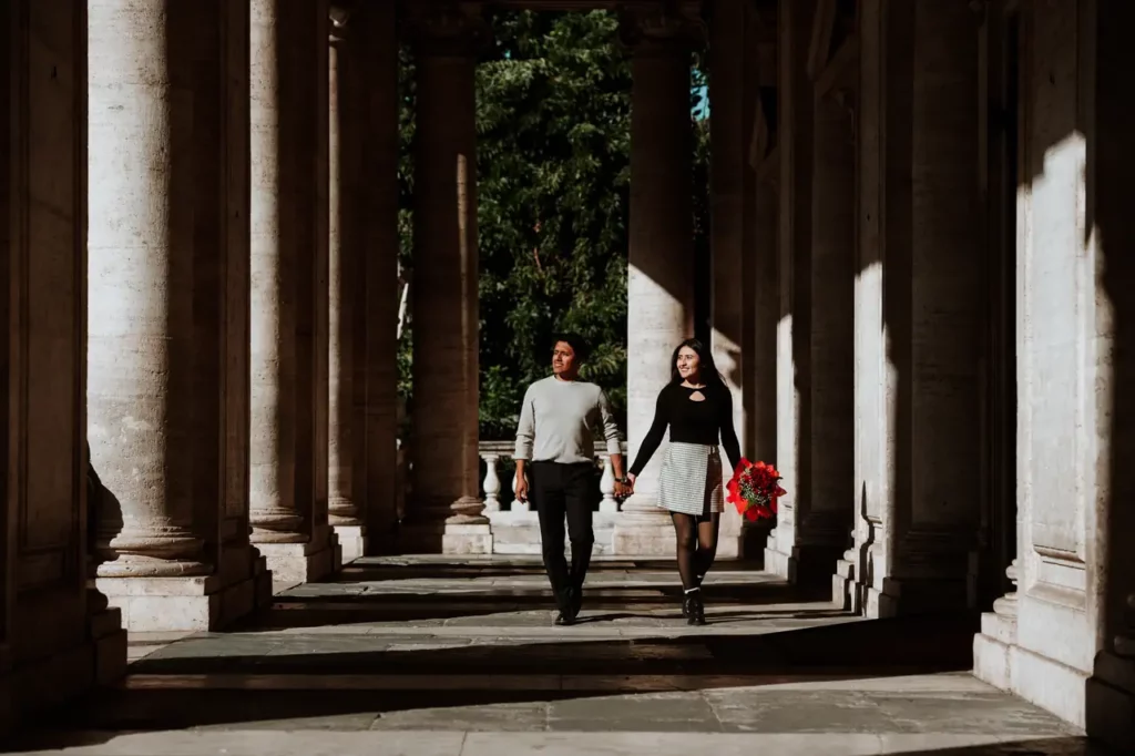 A couple holding hands and walking through a corridor with columns, with the woman carrying a bouquet of red flowers.