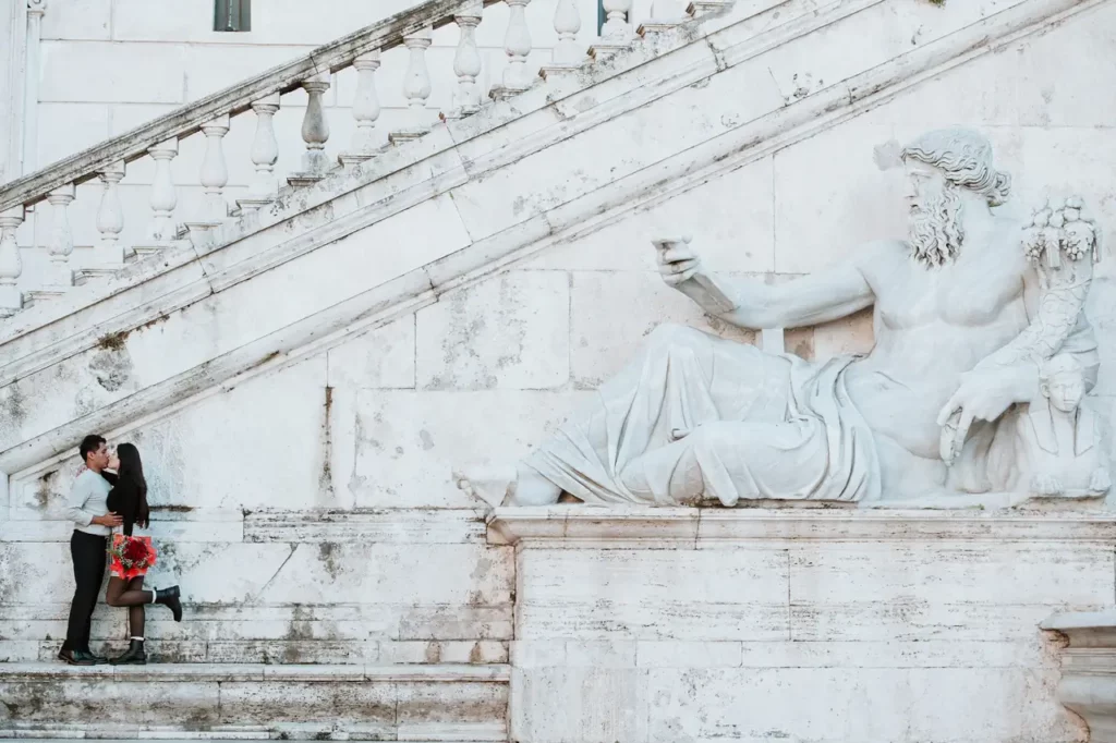 A couple kiss next to a large marble statue of a reclining bearded figure holding a book, located at the base of an ornate staircase.