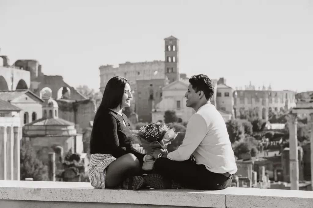 A couple sitting facing each other on a ledge, holding flowers, with historic buildings of rome in the background, in black and white.