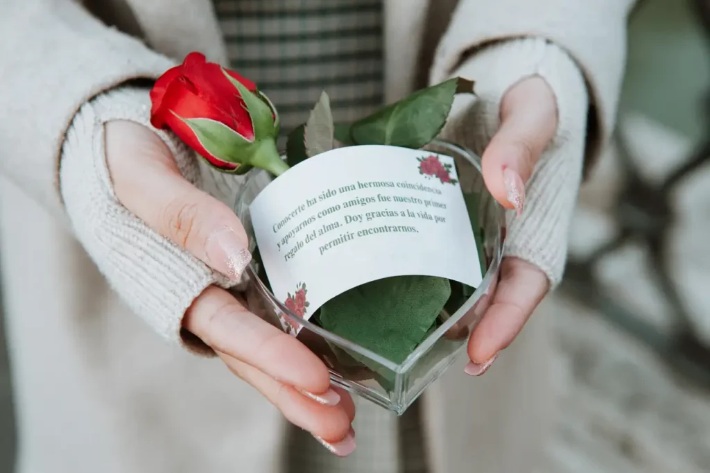 Hands holding a small red rose placed on green leaves inside a transparent heart-shaped container with a note wrapped around it.