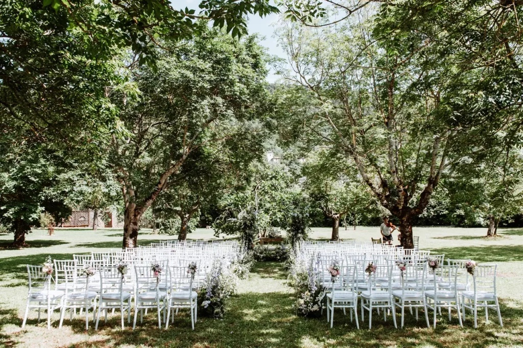 Set-up for outdoor wedding ceremony with white chairs arranged in rows, flanked by trees on either side and floral arrangements decorating the aisle.
