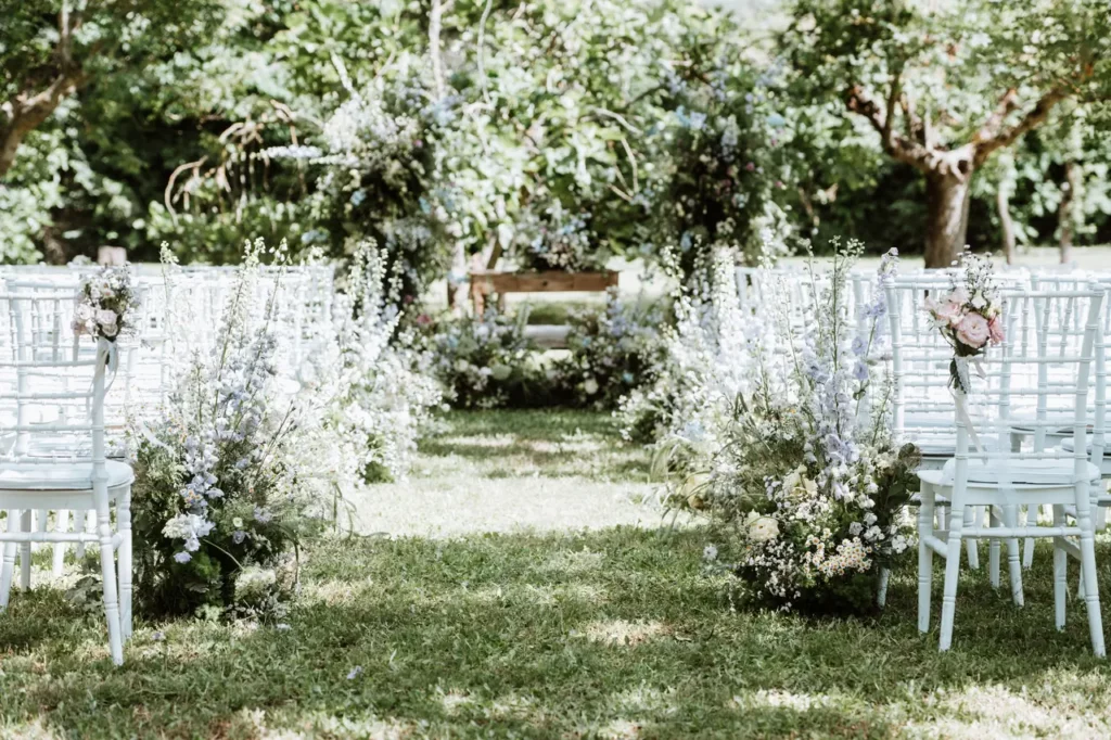 A set-up for an outdoor wedding ceremony with white chairs arranged in rows on a lawn, adorned with floral decorations. In the background is a wooden banquet with vegetation and flowers.