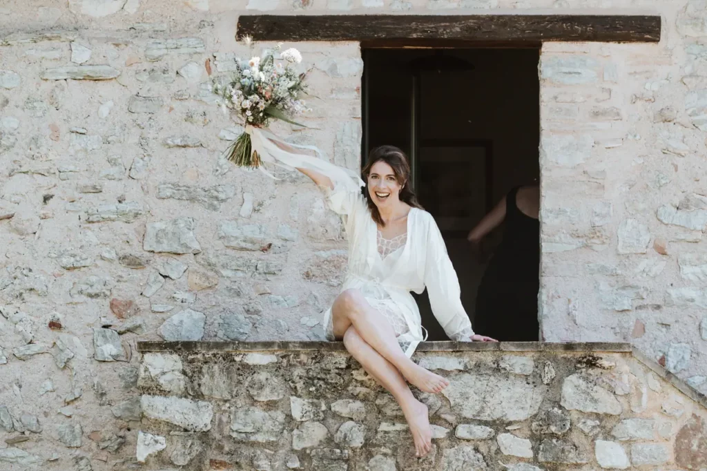 A woman dressed in white sits on a stone ledge in front of a stone wall, holds a bouquet of flowers above her head and smiles.