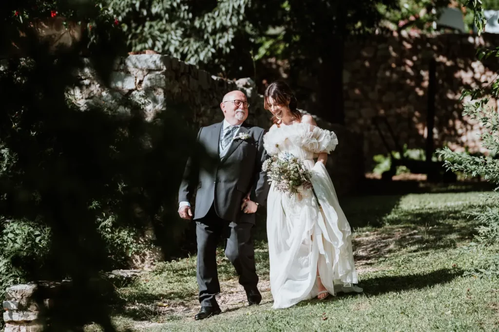 A bride in a white wedding dress walks outdoors holding hands with an elderly man in a suit. The bride and her father walk along a grassy path surrounded by trees and stone walls.