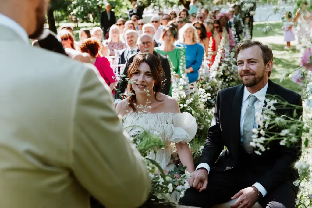 A bride and groom sit smiling before an officiant during an outdoor wedding ceremony, with guests seated behind them.
