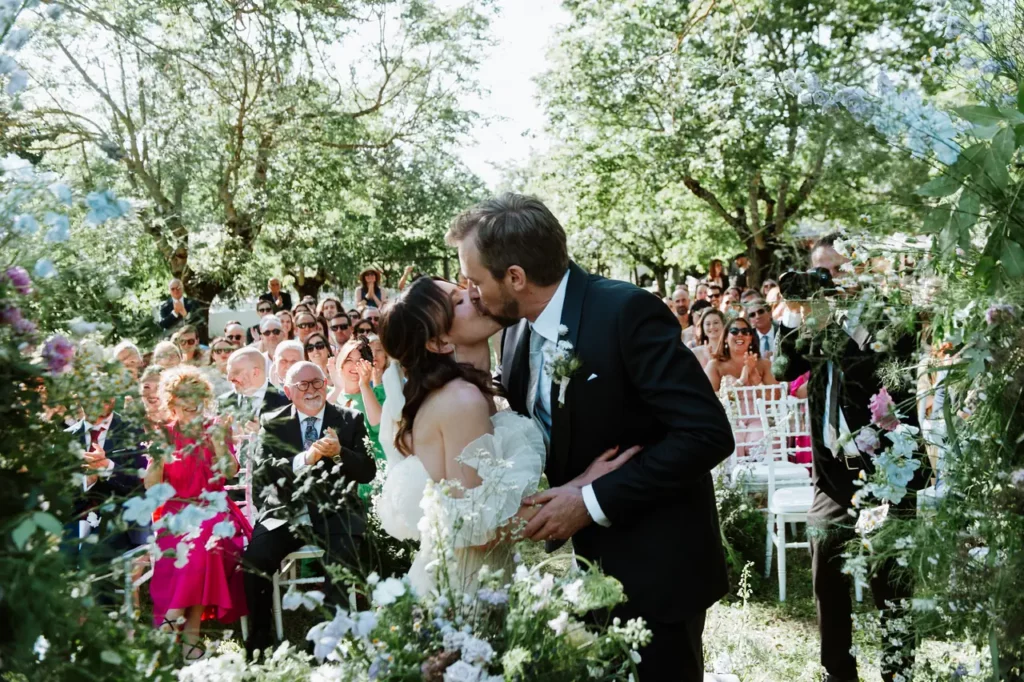 Bride and groom kiss during an outdoor wedding ceremony surrounded by guests seated in a garden with trees and flowers.