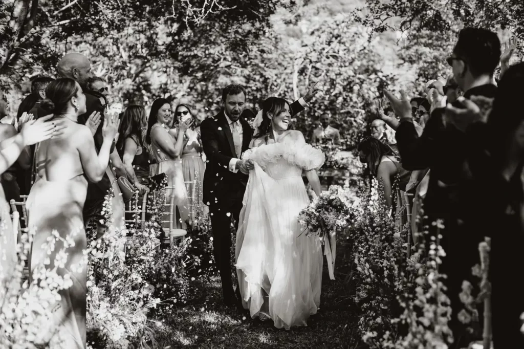 A bride and groom walk down an outdoor aisle as they are showered with confetti, surrounded by cheering guests. Garden flowers line the path. Black and white image.